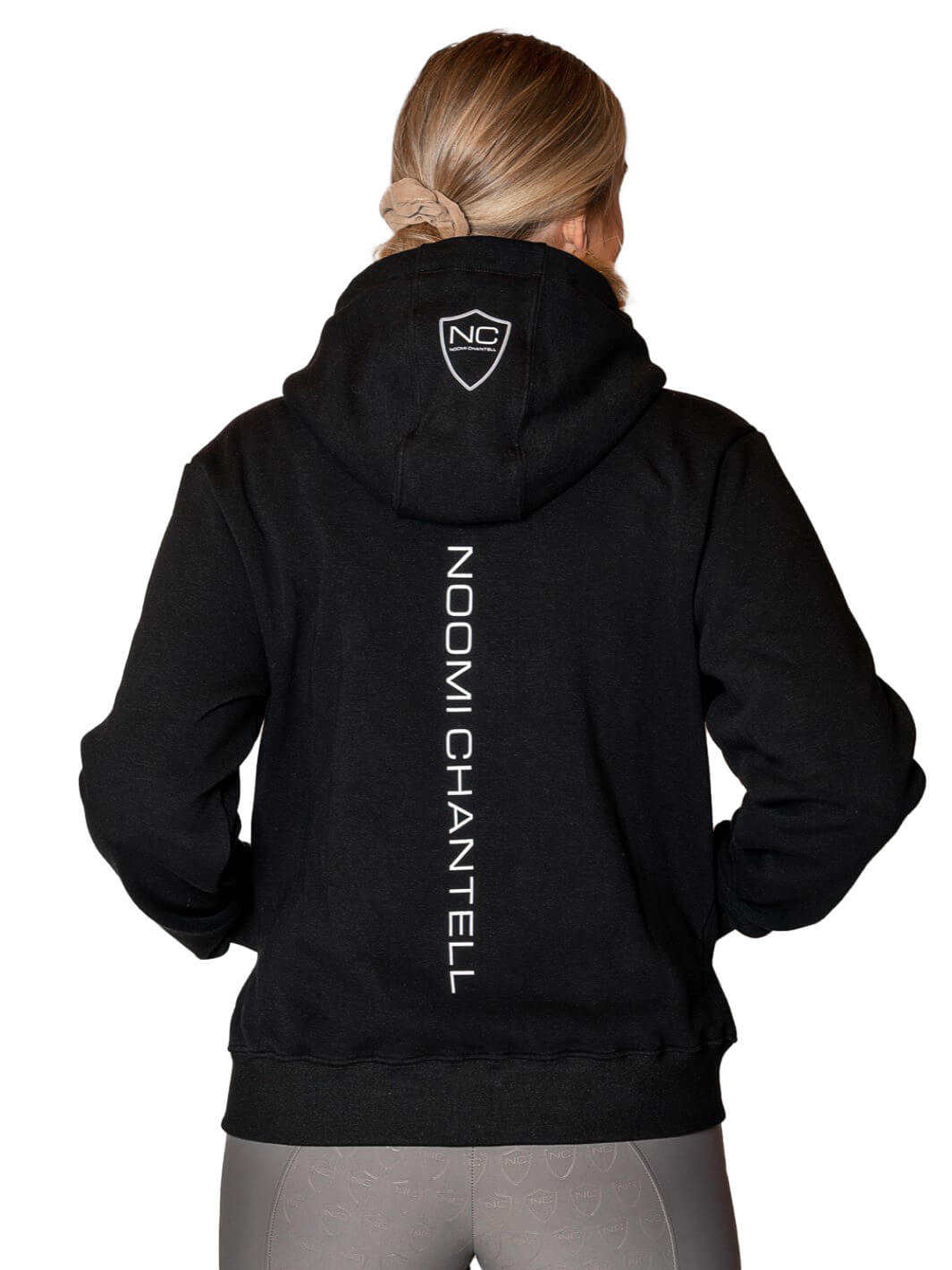 På bilden står modellen i en svart ridhoodie och visar ryggen av tröjan. Den är en vit stor textlogga Noomi Chantell uppifrån och ner på ryggen. Det är även en vit logga på luvan. Bakgrunden av bilden är vit